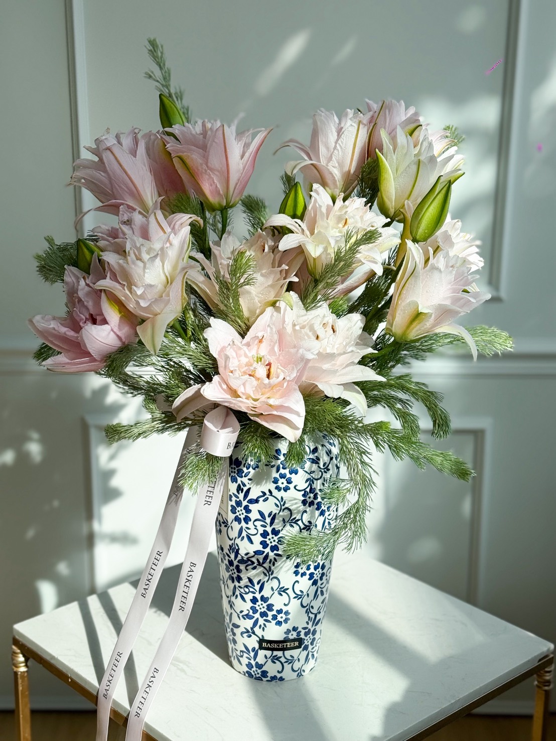 A blue and white patterned vase holds a bouquet of pale pink lilies and greenery, showcasing charming lily elegance. Adorned with a white ribbon, this artificial flower vase sits on a sunlit white table against a panelled wall.