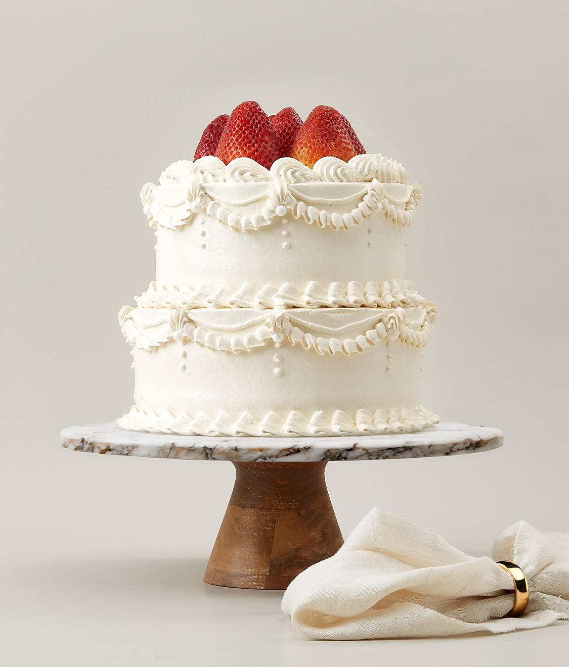 A two-tier white frosted cake topped with fresh strawberries is displayed on a white and brown marble cake stand. The cake is decorated with intricate piped designs and scalloped edges. A beige cloth with a golden ring lies nearby.