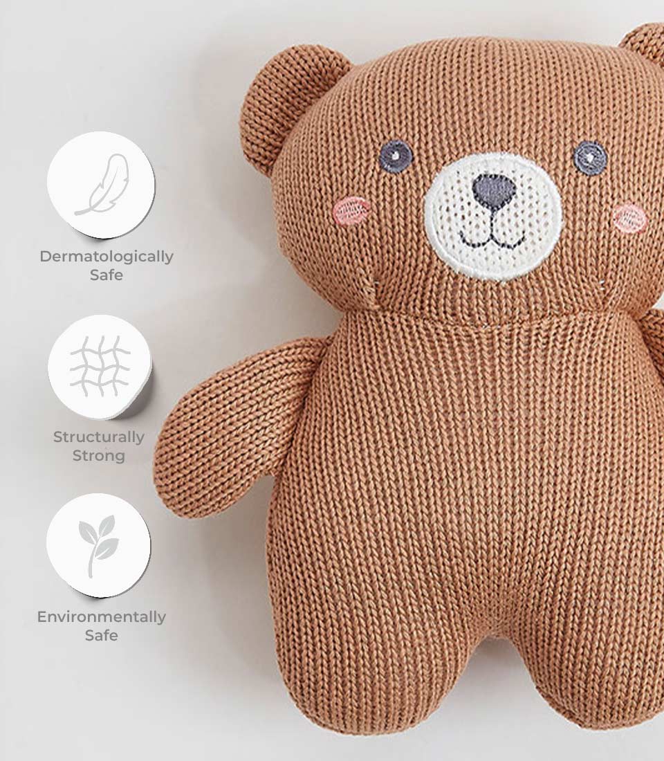 A knitted brown teddy bear with an embroidered face is positioned on a light background. Icons next to the bear illustrate that it is dermatologically safe, structurally strong, and environmentally safe.