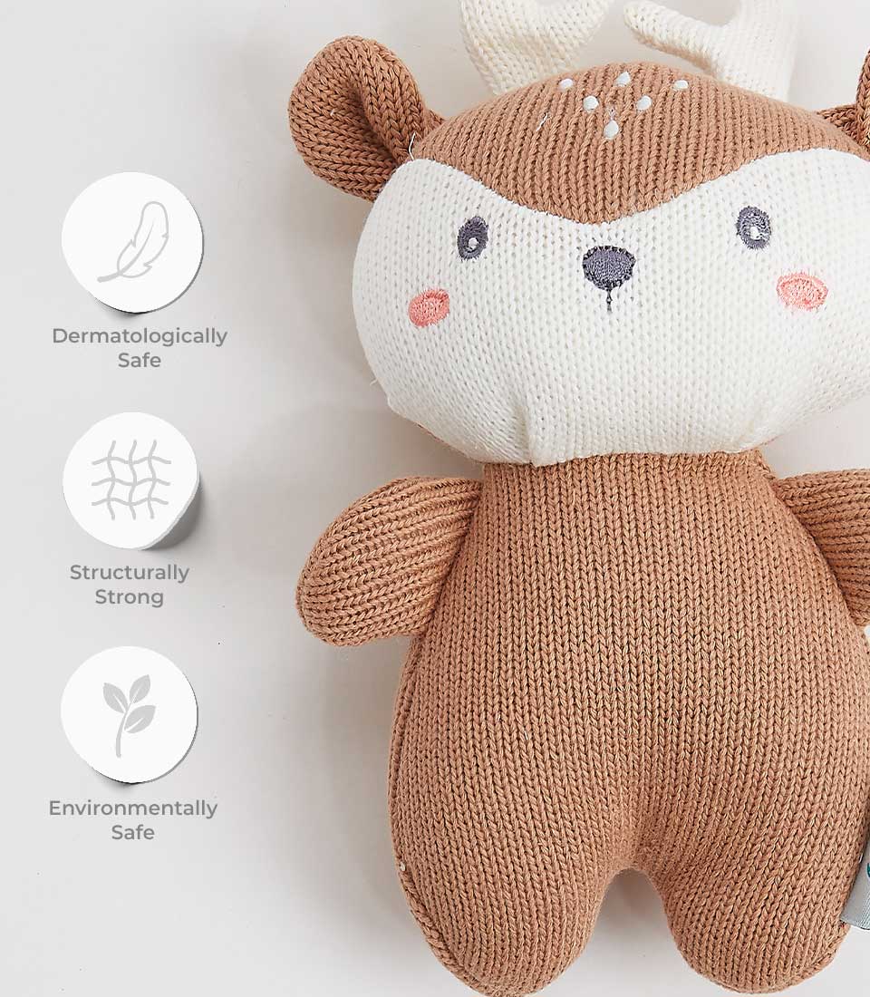 A knitted deer plush toy with a brown body and white face is shown against a light background. Next to the toy are three icons indicating that it is dermatologically safe, structurally strong, and environmentally safe.