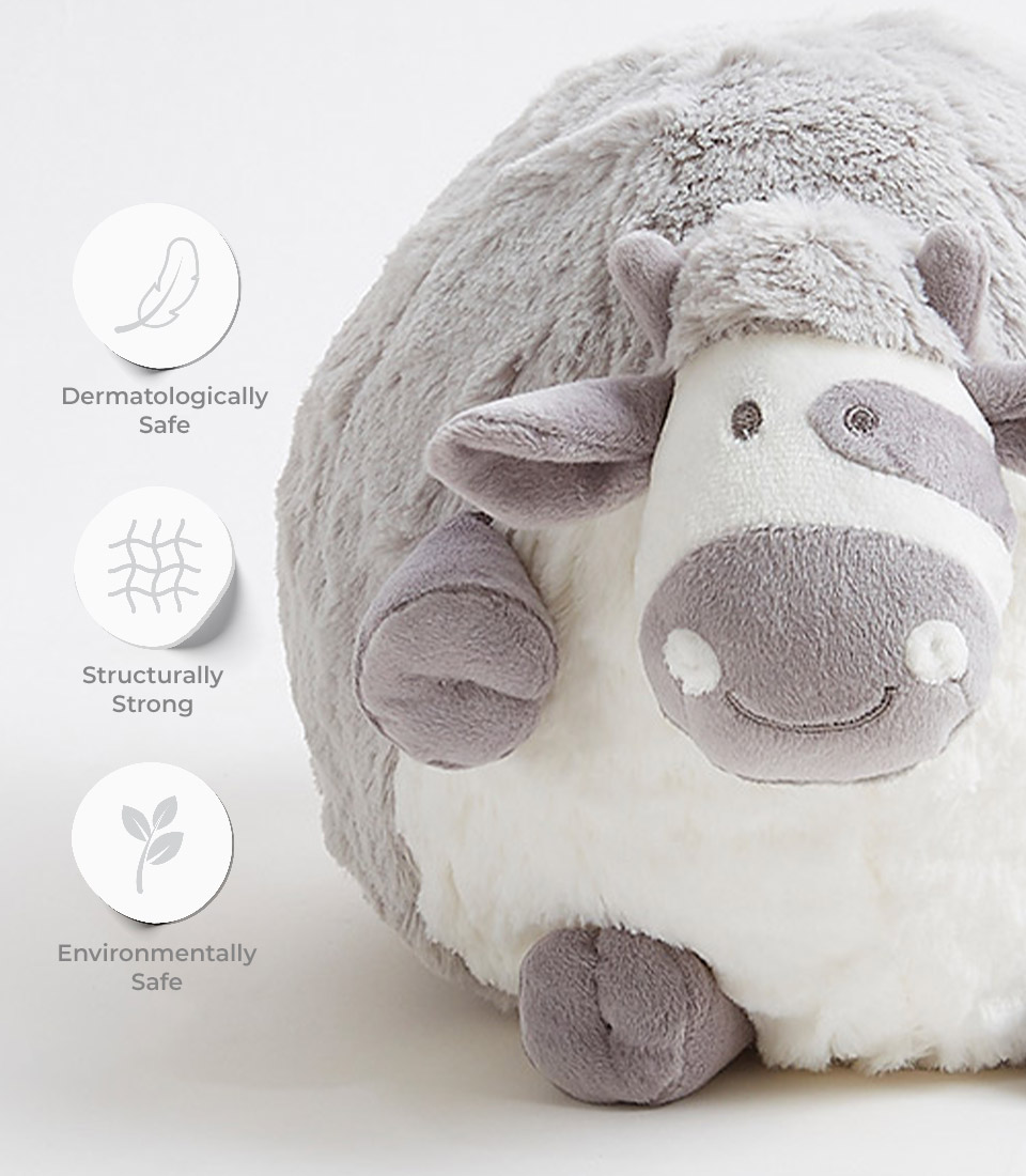 A chubby sheep soft toy with grey and white fur is shown. Three circular icons to the left highlight its features: 'Dermatologically Safe,' 'Structurally Strong,' and 'Environmentally Safe.' The sheep has a smiling face and soft, rounded body.