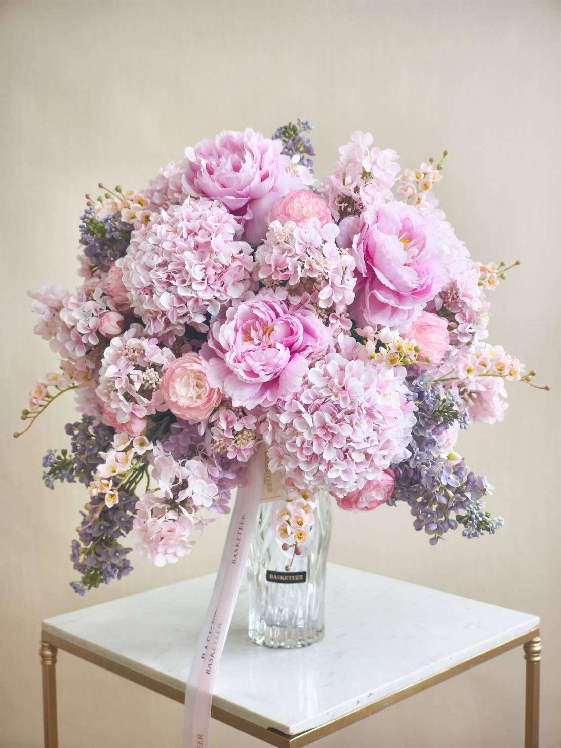 Elegant glass vase filled with pink peonies, soft pink hydrangeas, and lilac flowers, tied with a pastel pink ribbon.