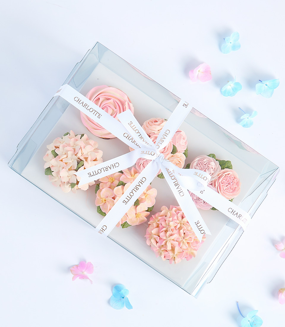 A clear box filled with pink and white cupcakes decorated with floral designs and ribbons.