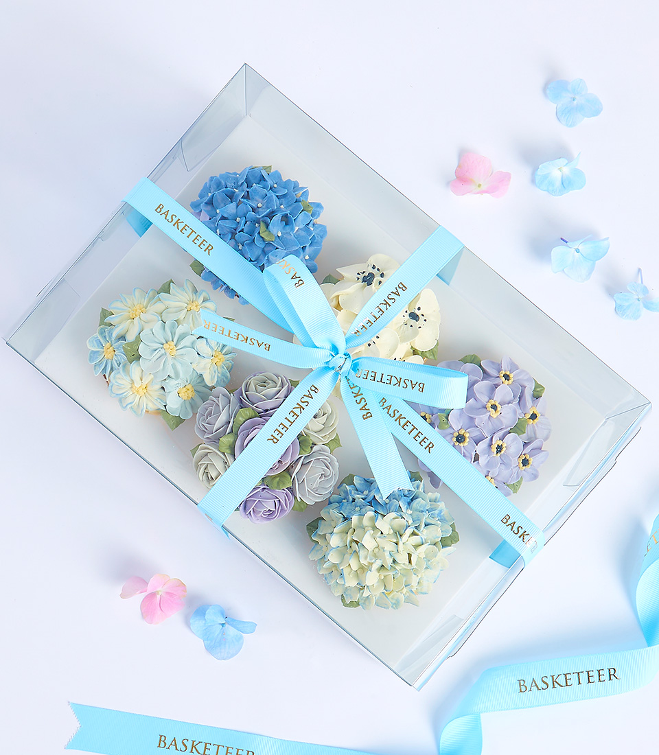 A clear box filled with cupcakes decorated with various floral designs in pastel colors, tied with a blue ribbon and adorned with the 