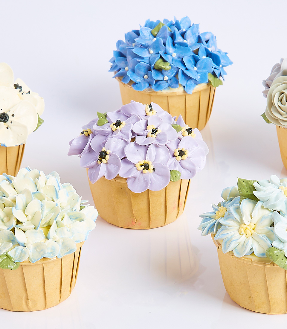 Assorted cupcakes decorated with intricate buttercream flowers in various pastel colors, resembling bouquets.