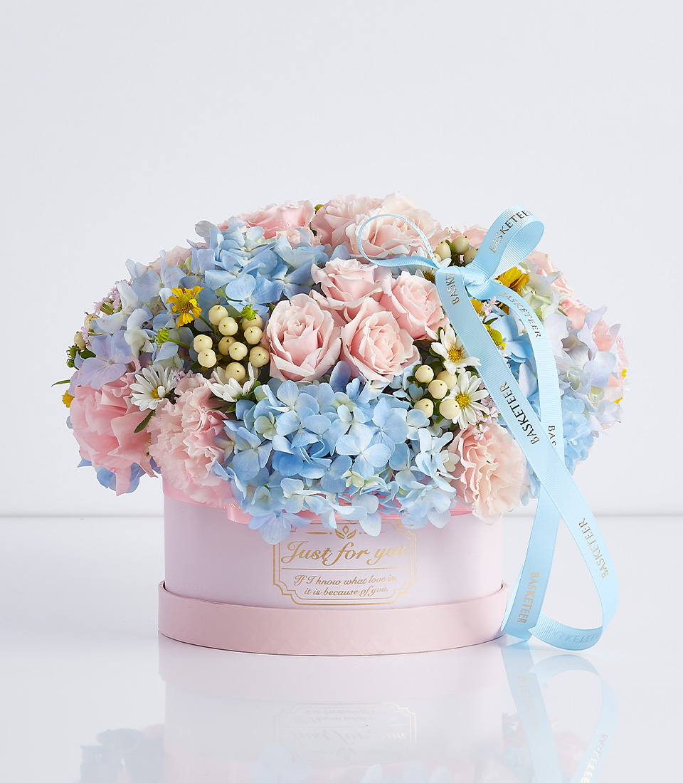 A pink hat box filled with pink roses, blue hydrangeas, white daisies, and yellow solidago, tied with a blue ribbon.