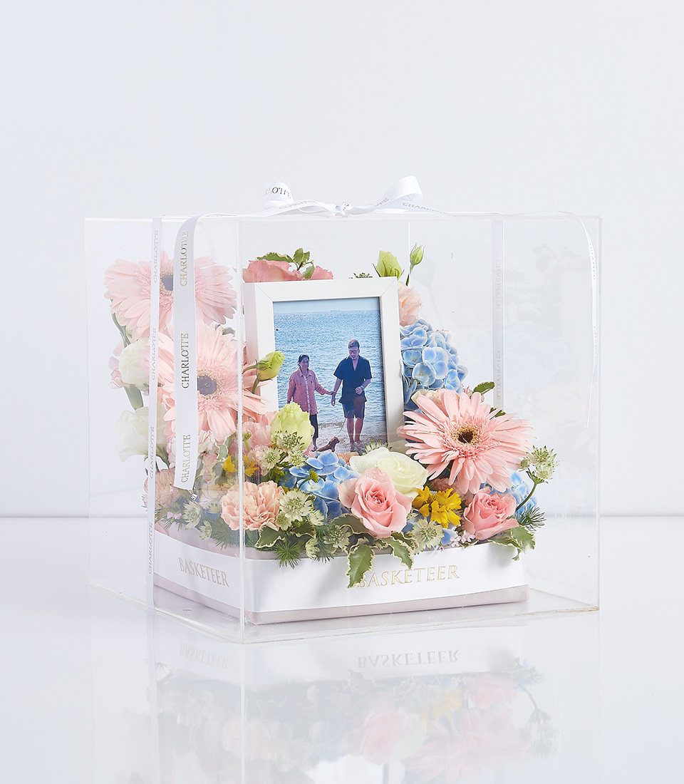 A clear gift box containing an arrangement of pink and blue flowers with a photo of two people walking on a beach. The box is decorated with ribbons labeled 