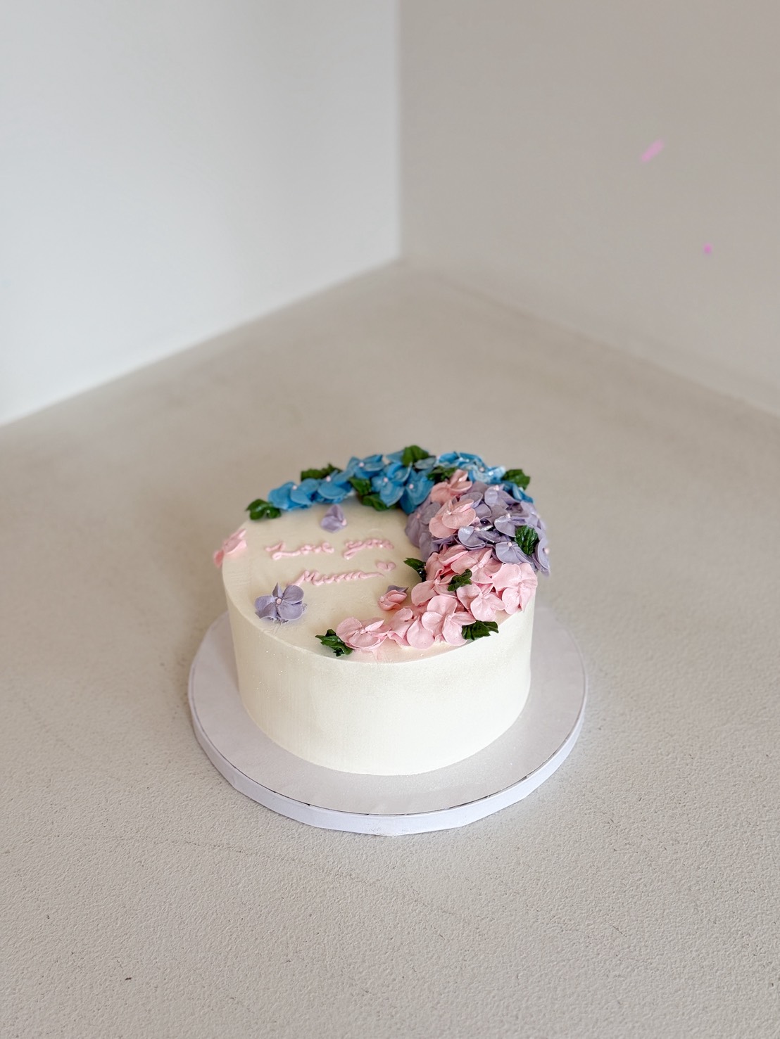 A round white Hydrangea Bloom Mother’s Day Cake decorated with pink, blue, and purple flower icing sits on a white cake board. Pink lettering is written on top. The cake rests on a light surface in a minimal corner setting.