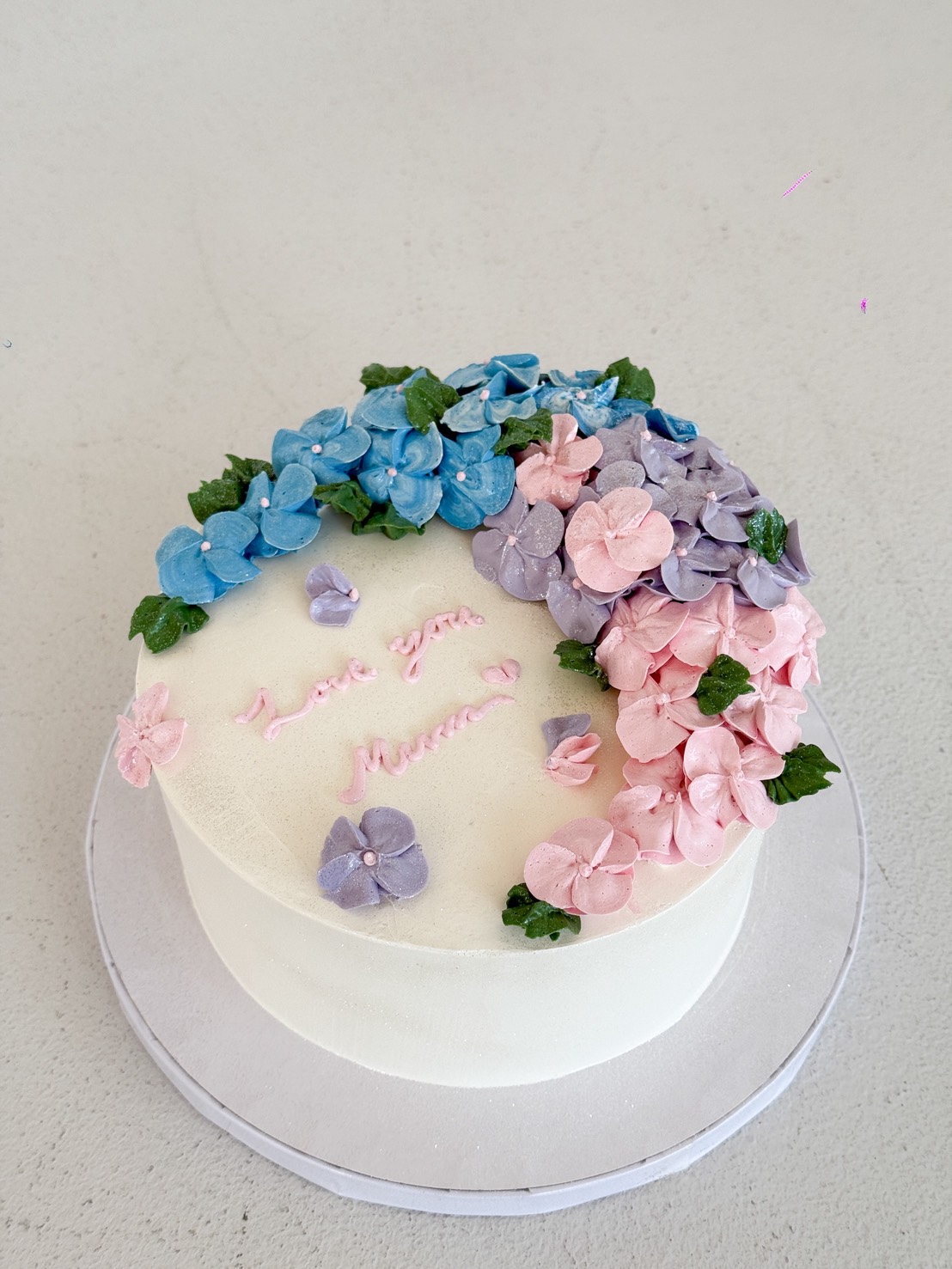 A round white Hydrangea Bloom Mother’s Day Cake adorned with pastel blue, pink, and purple hydrangea flowers and green leaves. 