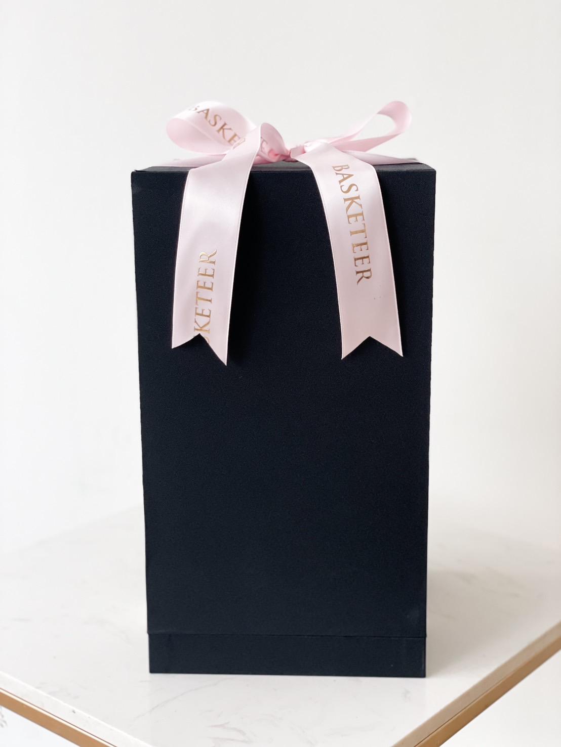 A tall, rectangular black gift box is adorned with a light pink ribbon that features the text 