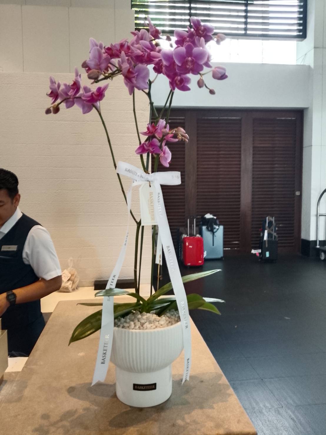 A white ceramic pot with Pure Elegance Orchid blooms and white ribbons stands on a counter in a modern foyer. A person in uniform is partially visible on the left, with luggage seen in the background.