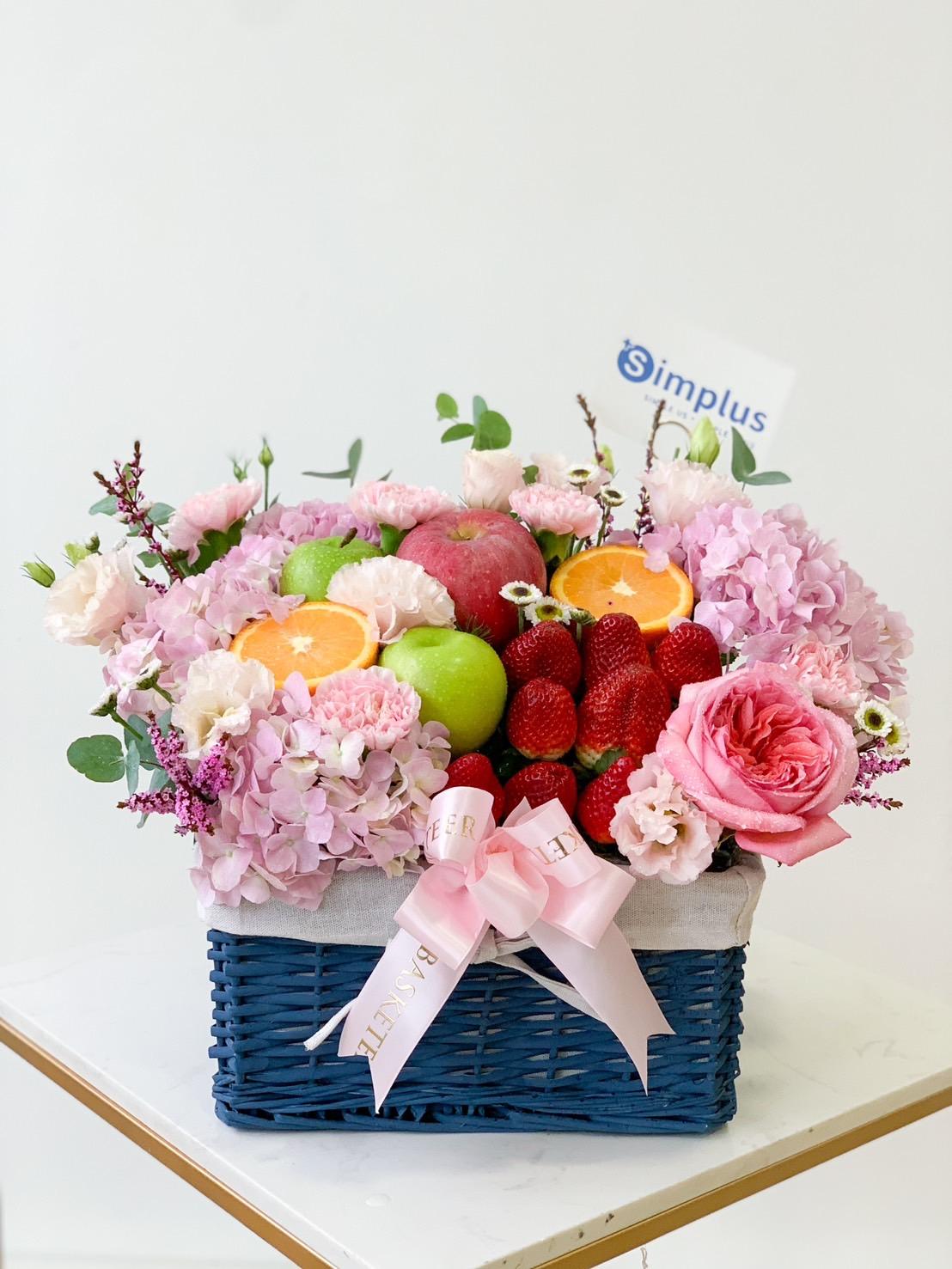 The Ocean Breeze Fruit & Floral Basket is a blue woven beauty filled with pink flowers, including roses and hydrangeas, accompanied by green and red apples, strawberries, orange slices, and a note card from 