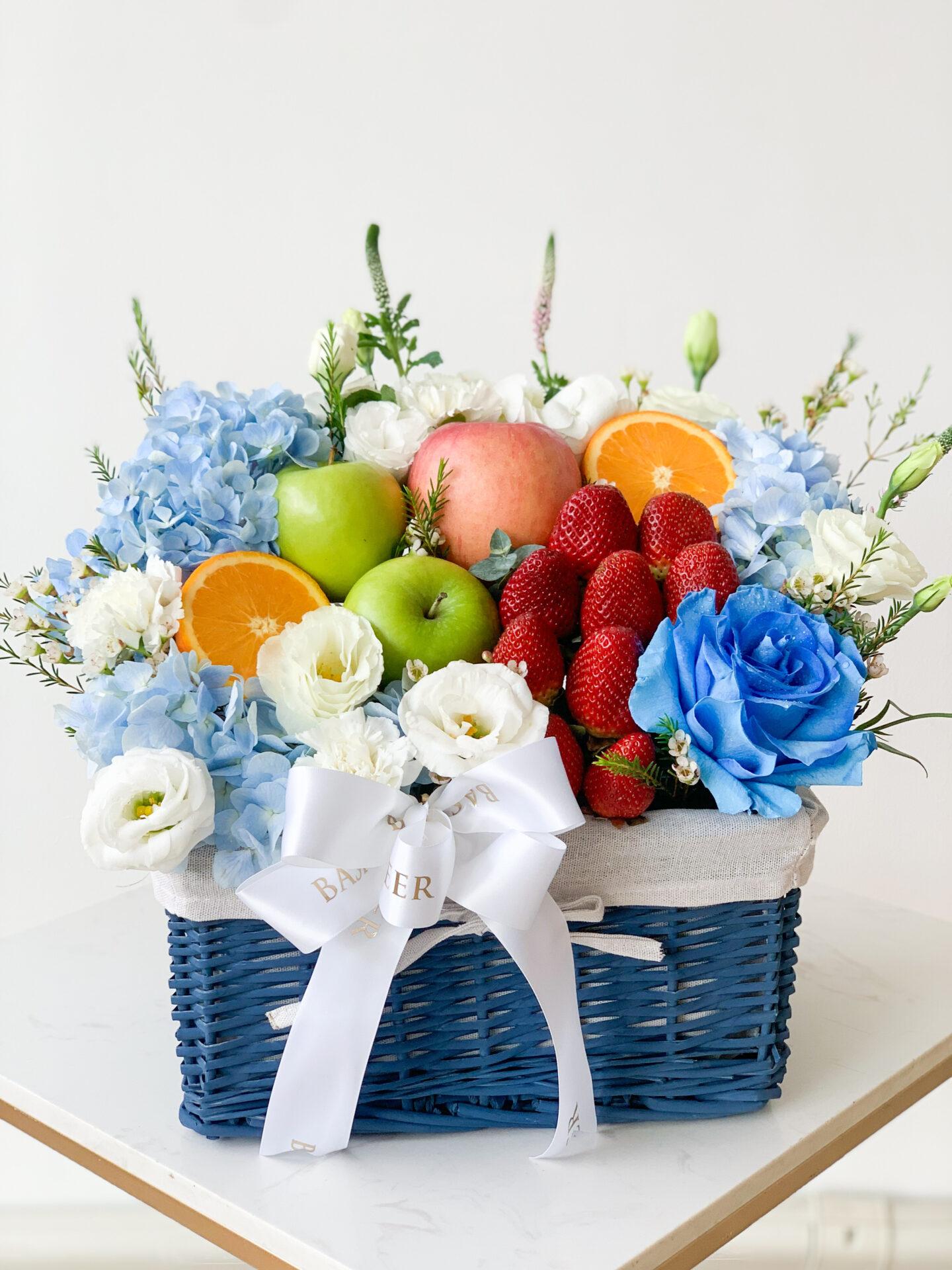 A vibrant Ocean Breeze Fruit & Floral Basket for Mother's Day filled with fresh fruits, including apples, oranges, and strawberries, surrounded by blue hydrangeas and white roses. A white ribbon bow with 
