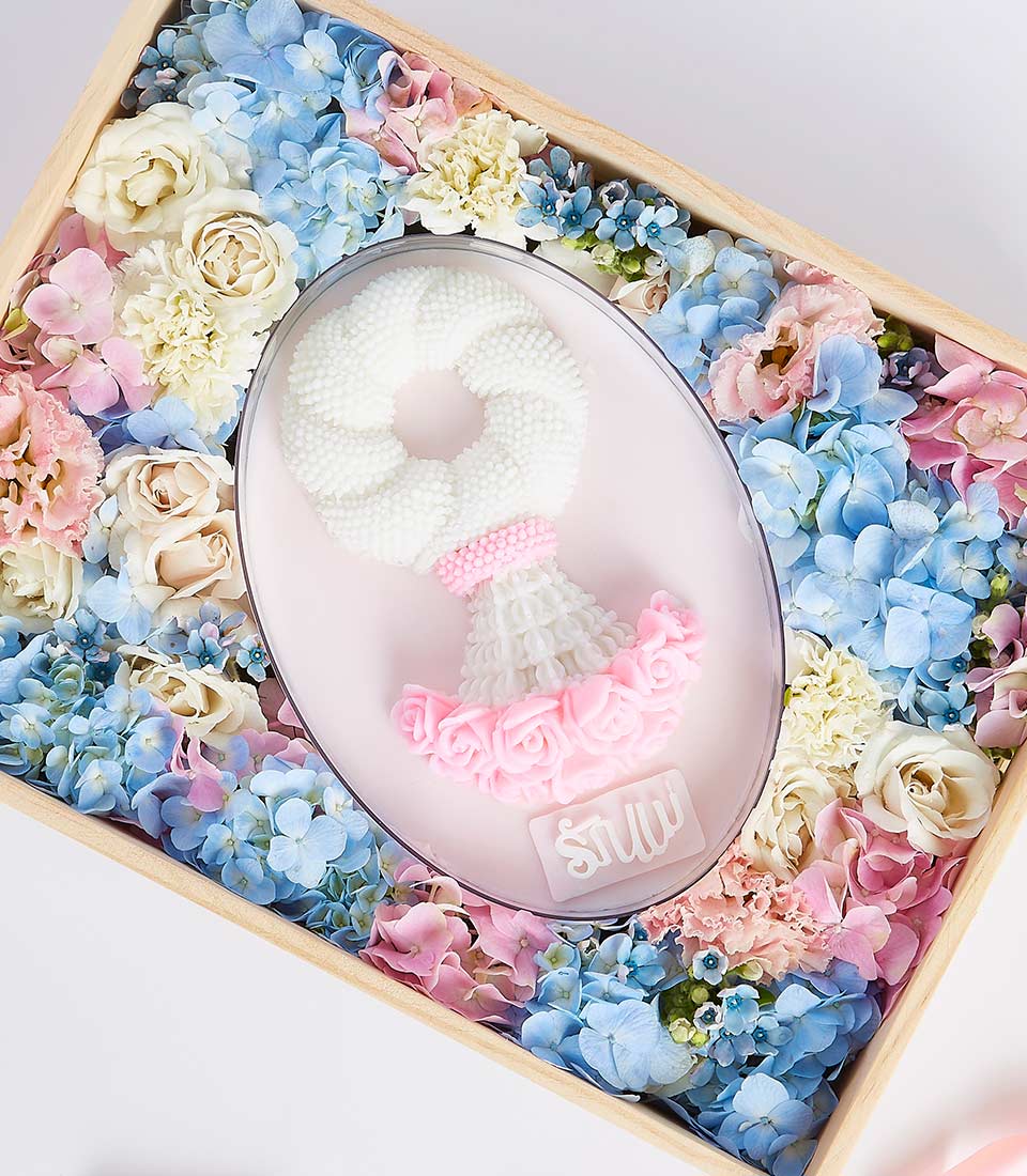 A delicate floral arrangement of pastel-colored flowers, including blue hydrangeas, pink and cream roses, surrounds a centerpiece white garland with pink accents in an oval wooden box. The garland is intricately crafted and rests on a white engraved plaque—perfect as a Jelly Malai for Mother's Day gift.