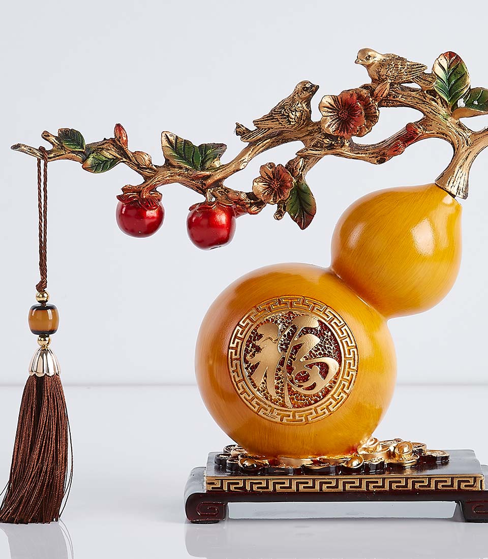 Golden gourd figurine with a curved stem holding two birds and red berries. The gourd has intricate carvings and rests on a black wooden base with a tassel.