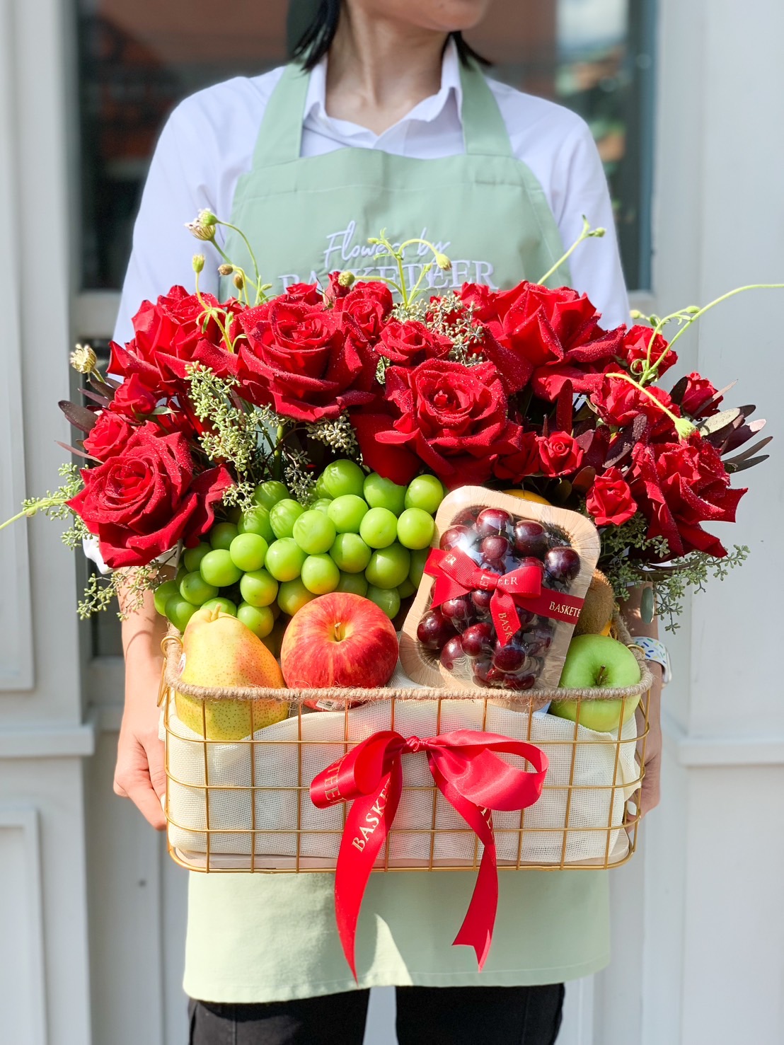 A person wearing a green apron holds a Mother's Day Flower and Fruit Basket filled with vivid red roses and various fruits, including green grapes, apples, and a pack of cherries. The decorative basket is adorned with red ribbons.
