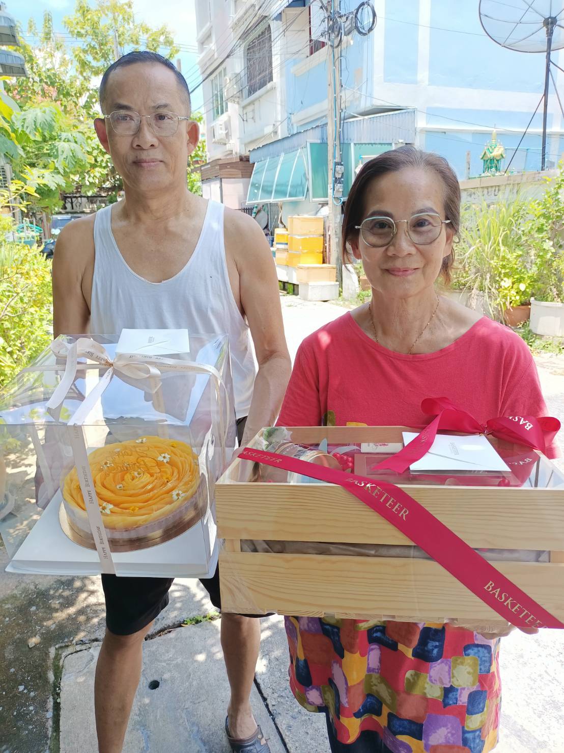 A man and a woman are standing outside holding gifts. The man on the left is wearing a white tank top and glasses, holding a cake in a clear box. The woman on the right is wearing a red shirt and glasses, holding a wooden gift box with a red ribbon.
