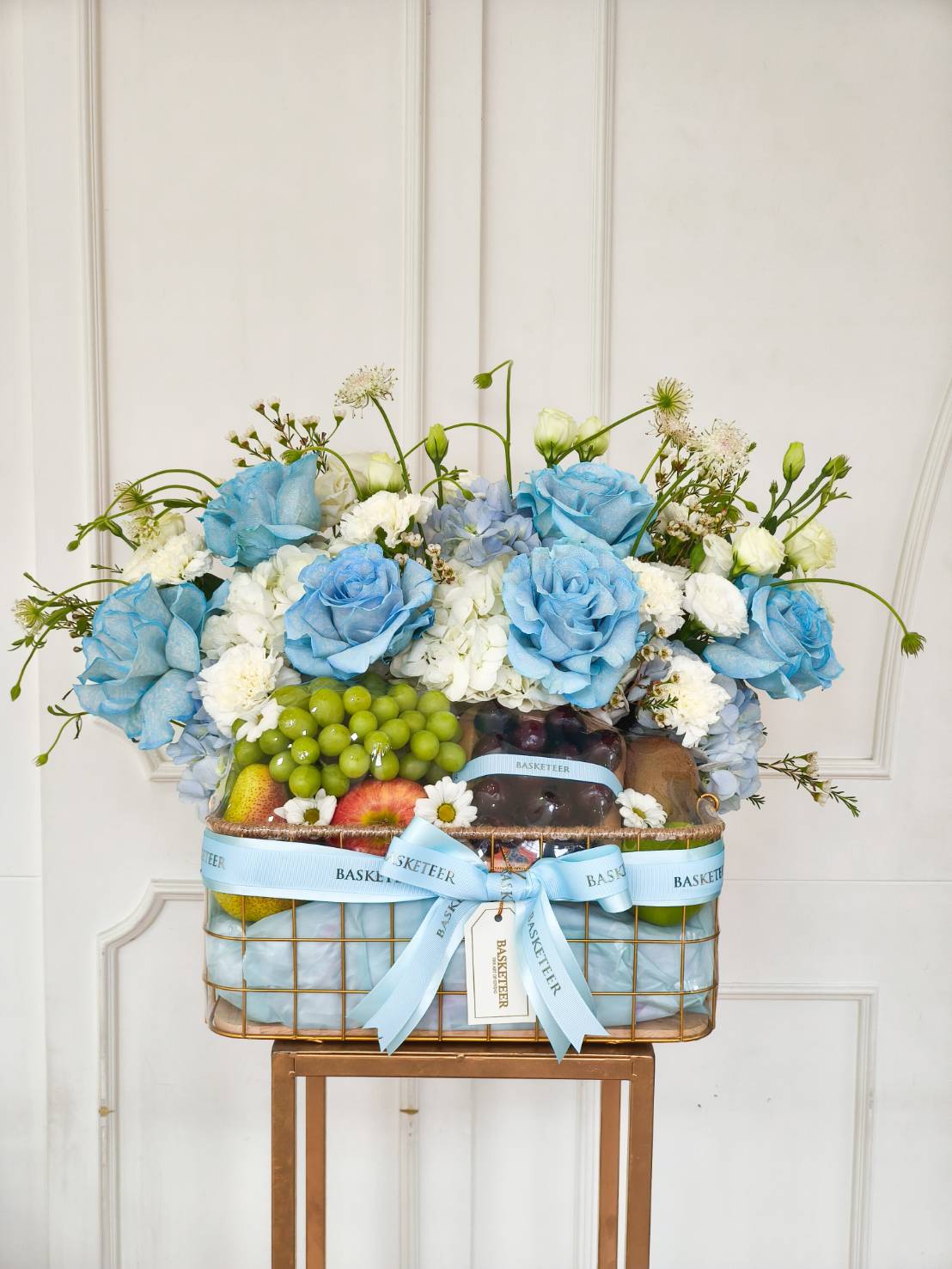 Luxury fruit and flower gift basket with blue roses, white hydrangeas, green grapes, apples, pears, and other fresh fruits, tied with a blue Basketeer ribbon.
