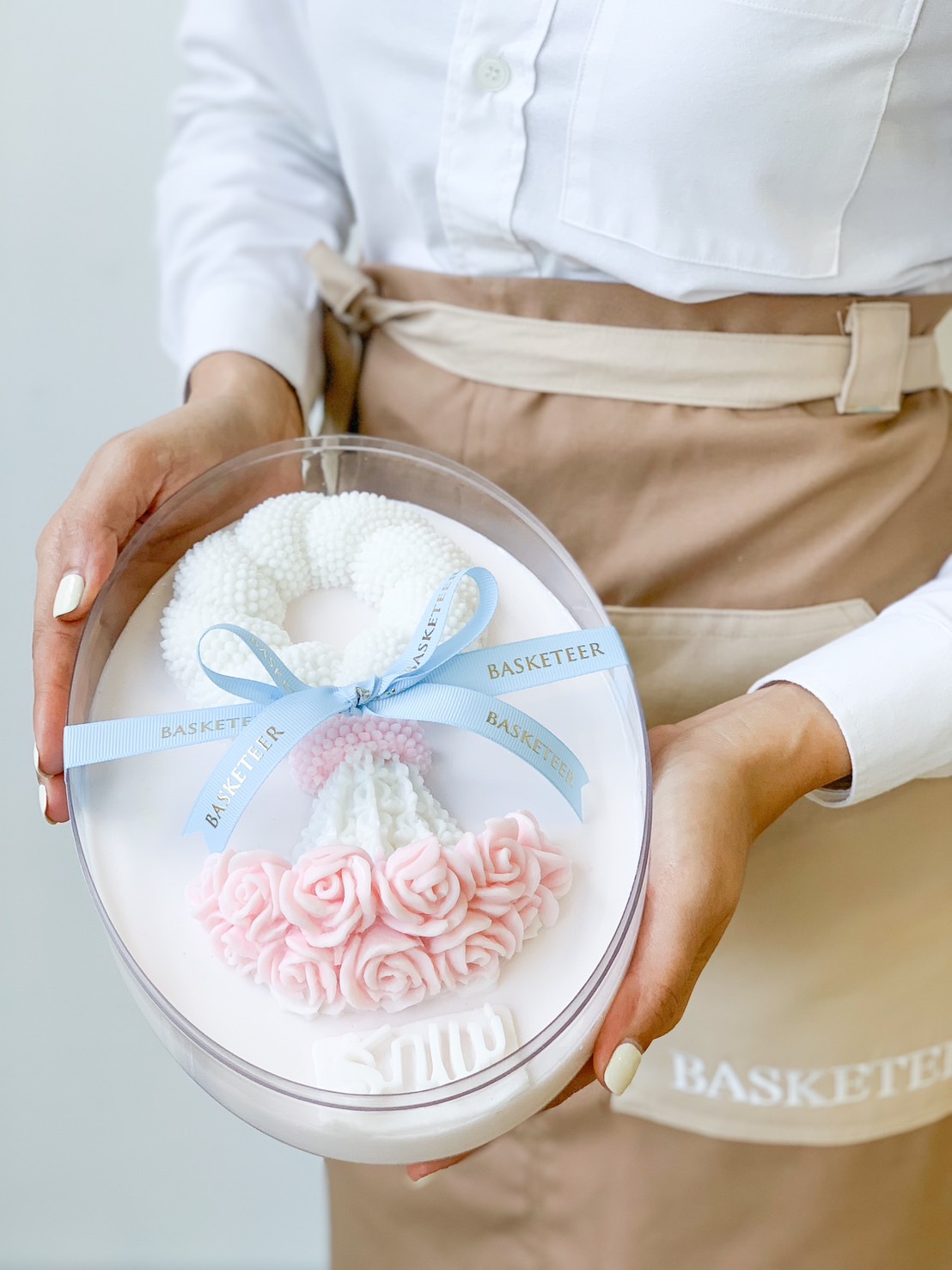 A person in a beige apron holds a clear oval gift box containing white and pink decorative floral soap pieces tied with a blue ribbon labeled 