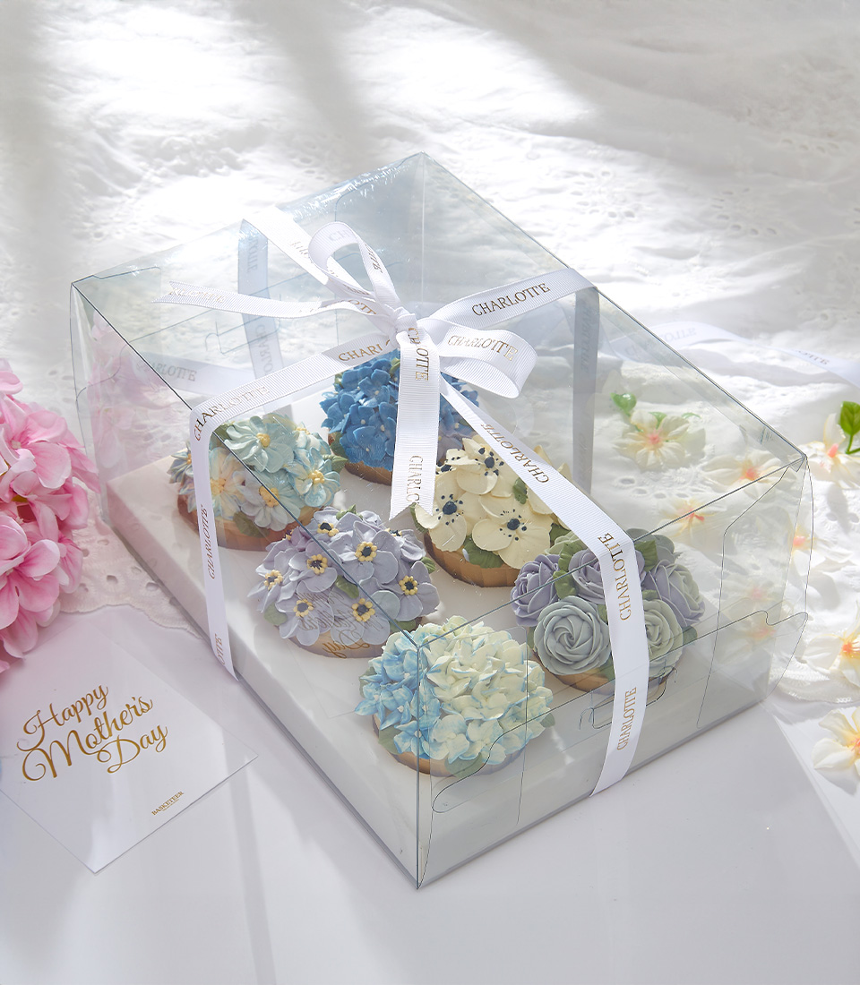 A set of Serenity Blossom Cupcakes with pastel-colored floral designs is neatly arranged in a clear box tied with a white ribbon. The box is placed on a white tablecloth with pink flowers and a 