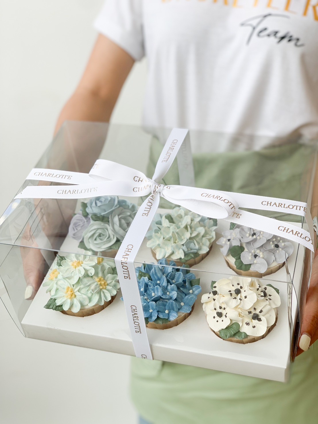 A person holding a clear box containing six intricately decorated floral cupcakes. The Serenity Blossom Cupcakes feature designs of blue, white, and yellow flowers with green leaves. The box is tied with a white ribbon that has 