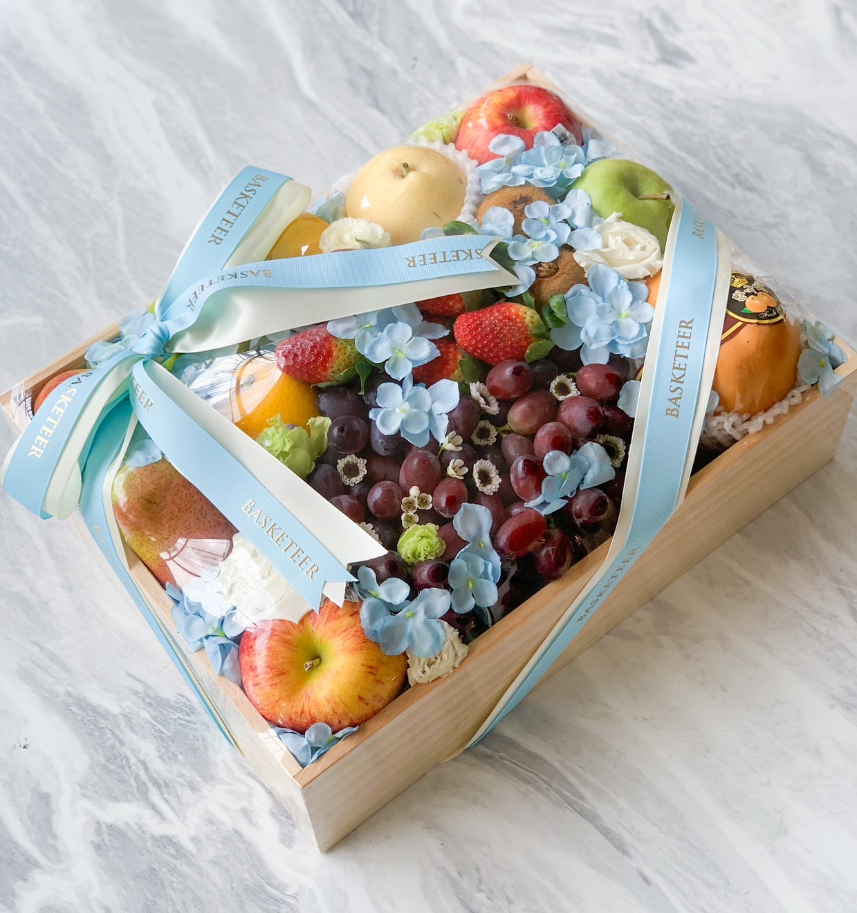 A bountiful Mother's Day wooden box filled with an assortment of fruits, such as apples, grapes, and a pear, covered with blue ribbon and decorated with light blue flowers. The box is tied with a bow made from ribbon labeled 