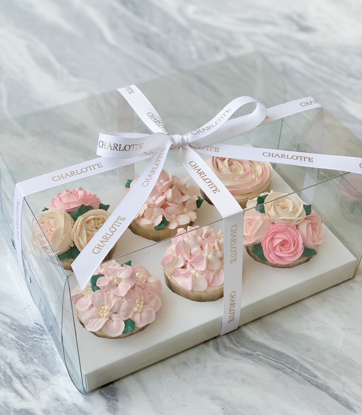 A clear gift box containing six beautifully decorated Blossom Bouquet Cupcakes sits on a marble surface. The cupcakes feature intricate floral designs in pink and cream colors, resembling roses and hydrangeas. The box is tied with a white ribbon printed with the name 