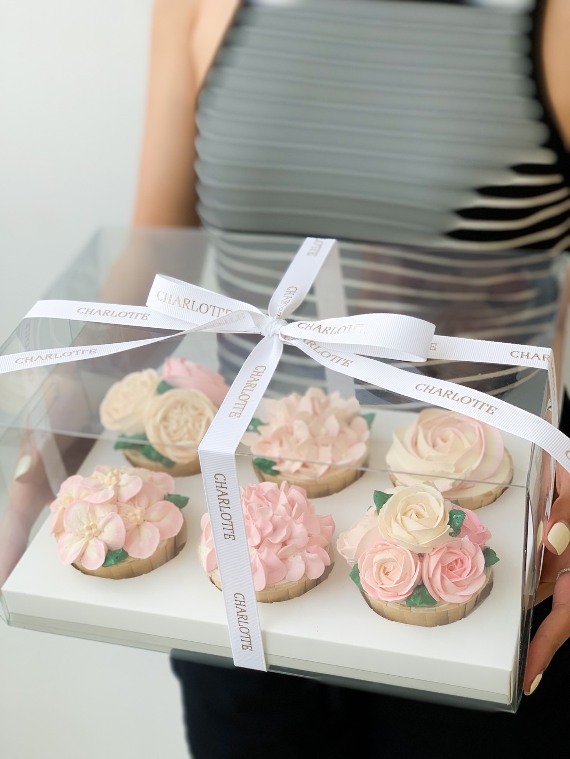 A person holding a transparent box containing six beautifully decorated Blossom Bouquet Cupcakes. The cupcakes are adorned with intricate pink and white flower designs, resembling roses and blossoms. The box is secured with a white ribbon that reads 