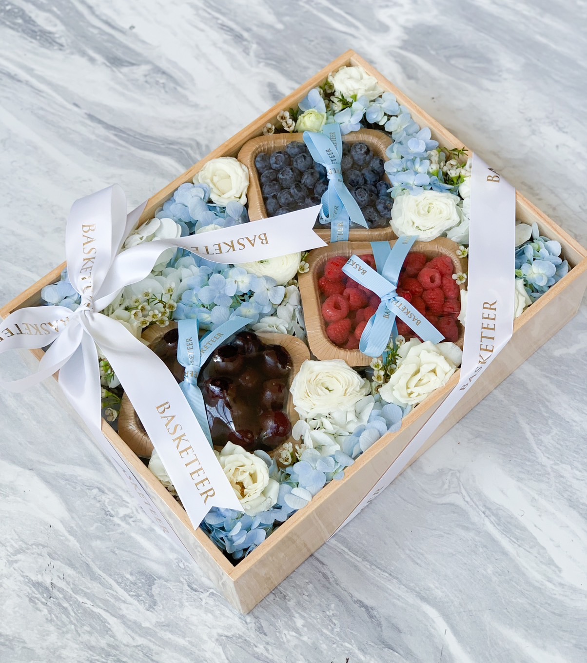 A wooden gift box with compartments contains fresh blueberries, raspberries, and dark cherries, beautifully decorated with white roses and blue hydrangeas. The compartments are tied with white ribbons labeled 