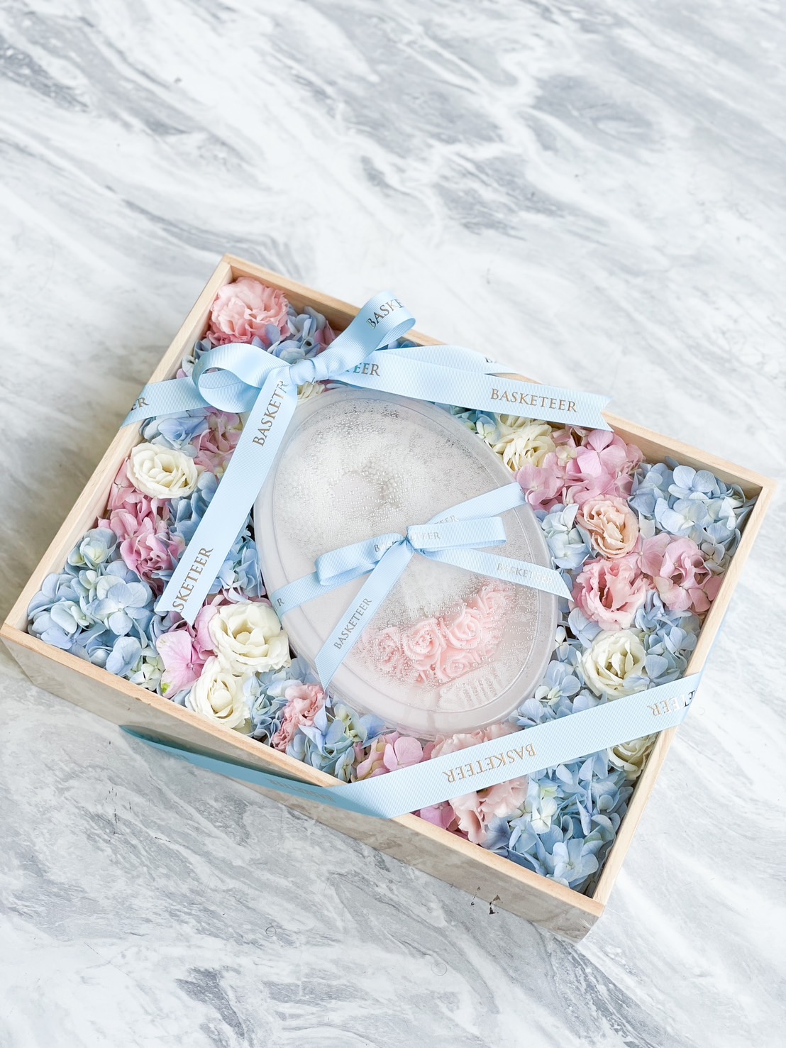A wooden gift box filled with pastel-colored flowers, including pink, blue, and white blossoms. In the center is a translucent shell-shaped container adorned with a blue ribbon labeled 