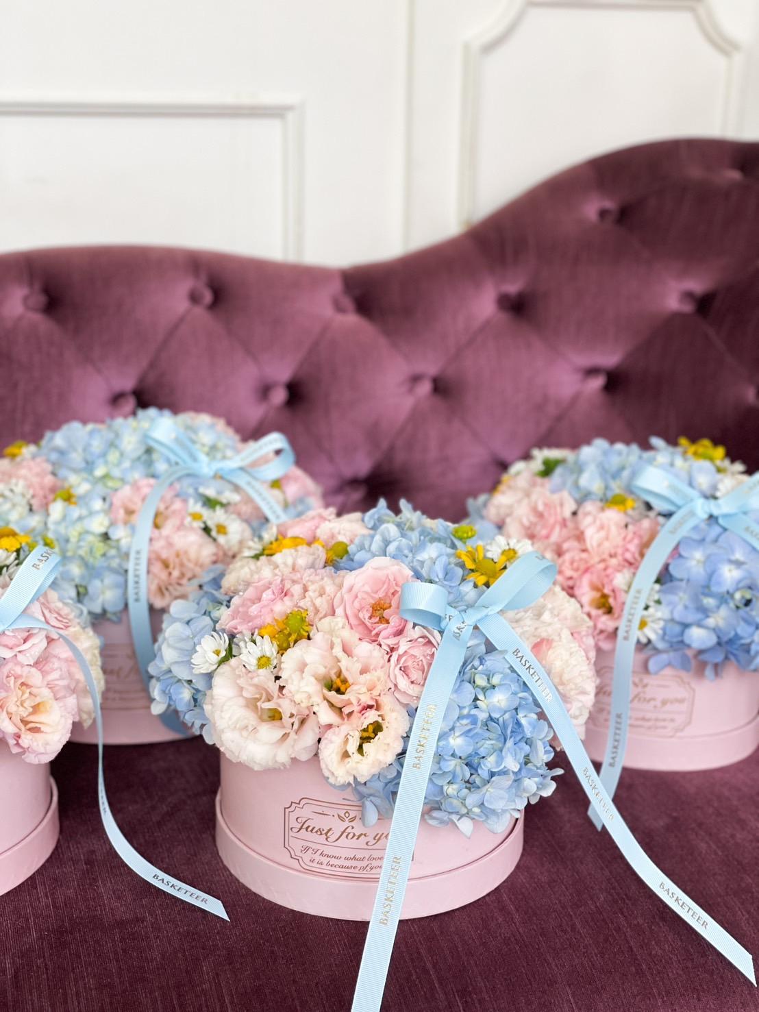 Three round blue and pink floral arrangements, including roses and hydrangeas, are placed on a plush mauve sofa. Each arrangement is adorned with a light blue ribbon. The backdrop features elegant white paneling.