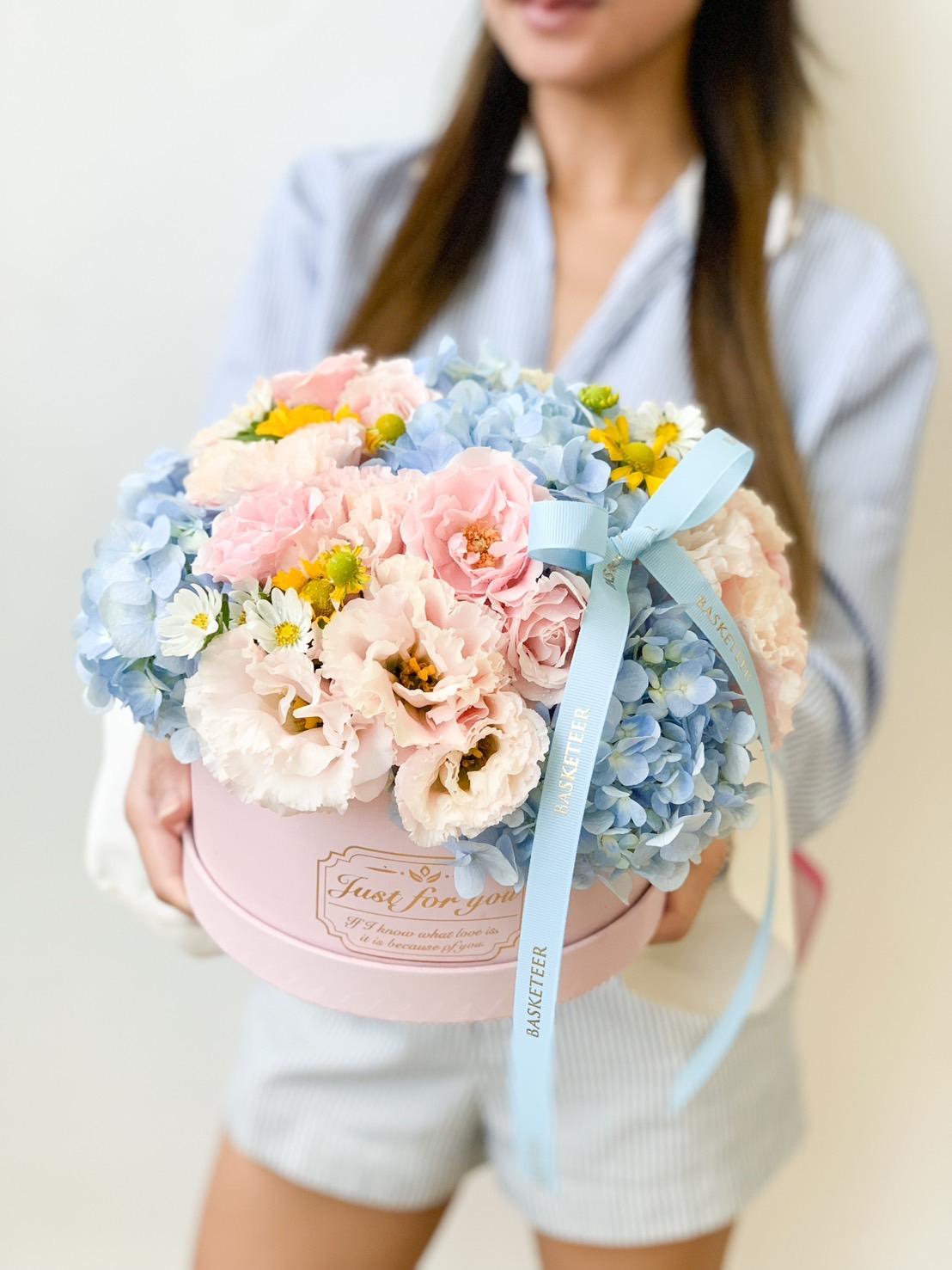 A woman in a light blue striped shirt holds a round pink floral arrangement featuring blue hydrangeas, pink roses, white flowers, yellow accents, and a light blue ribbon with the word 