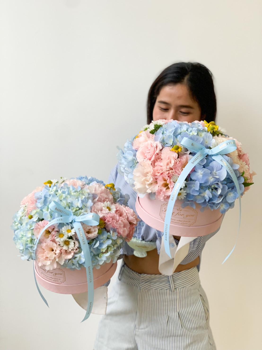 A person in a striped shirt holds two large round bouquets of flowers. The blue and pink floral arrangements, displayed in pastel blue and pink hat boxes, consist of blue, pink, and yellow blooms adorned with pastel blue ribbons. The background is a plain, light-colored wall.