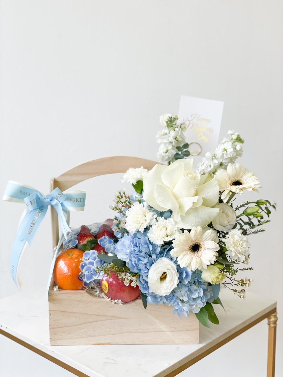 A wooden gift basket filled with blue and white flowers, including hydrangeas and gerberas, exudes a sense of freshness for Mother's Day. Accompanied by fresh fruits like oranges and pomegranates, the basket is adorned with a blue ribbon and a card, all set on a white marble surface.