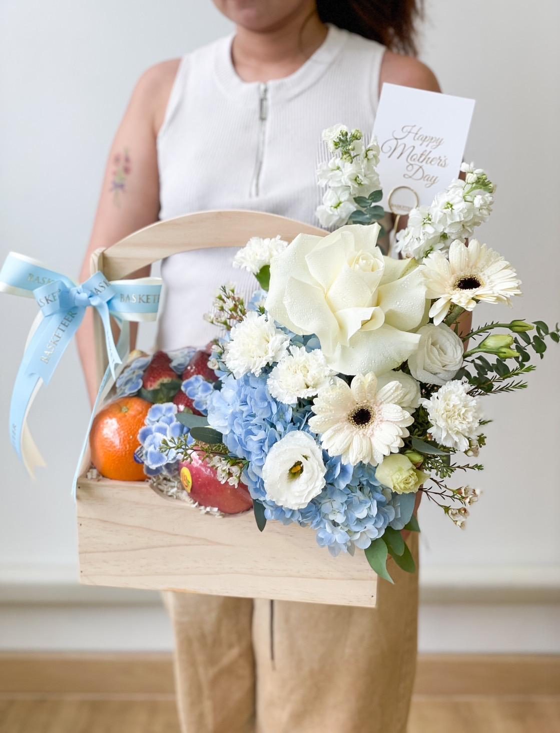 A person in a white sleeveless top holds a wooden crate filled with assorted fruits and a bouquet of flowers, including blue hydrangeas, white roses, and daisies. A card with 