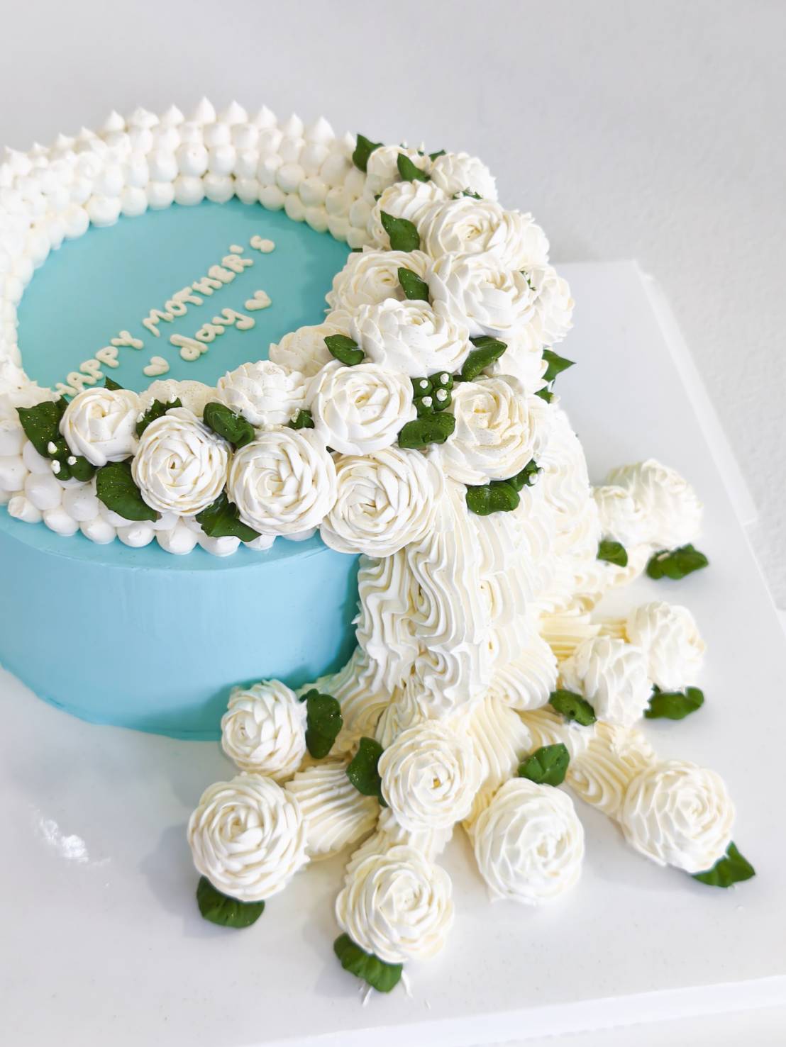 Blue Mother’s Day cake with white buttercream roses and garland design, featuring “Happy Mother’s Day” message.