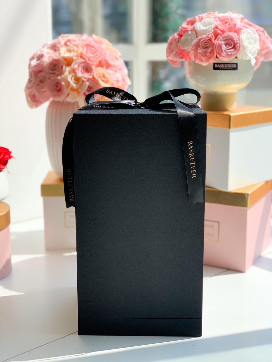 A luxury black gift box with a satin Basketeer ribbon, placed in front of elegant preserved rose arrangements in soft pastel tones.
