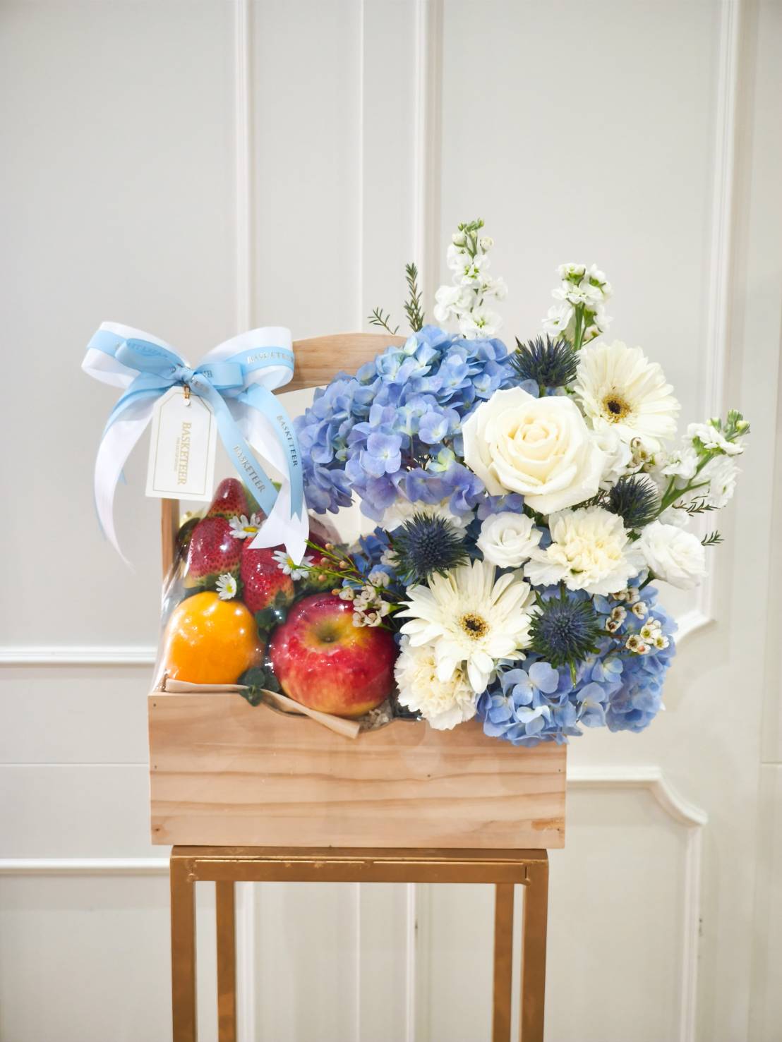 Wooden crate filled with fresh apples, oranges, and strawberries, decorated with blue hydrangeas, white roses, and gerberas.