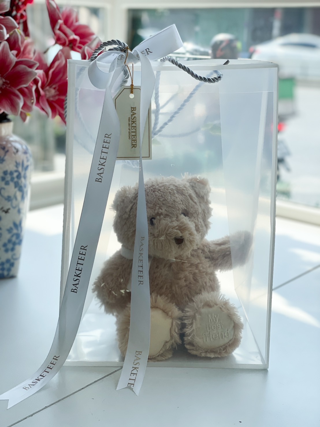 A soft, brown plush teddy bear with an embroidered message, elegantly packaged in a transparent gift bag with white satin ribbons.
