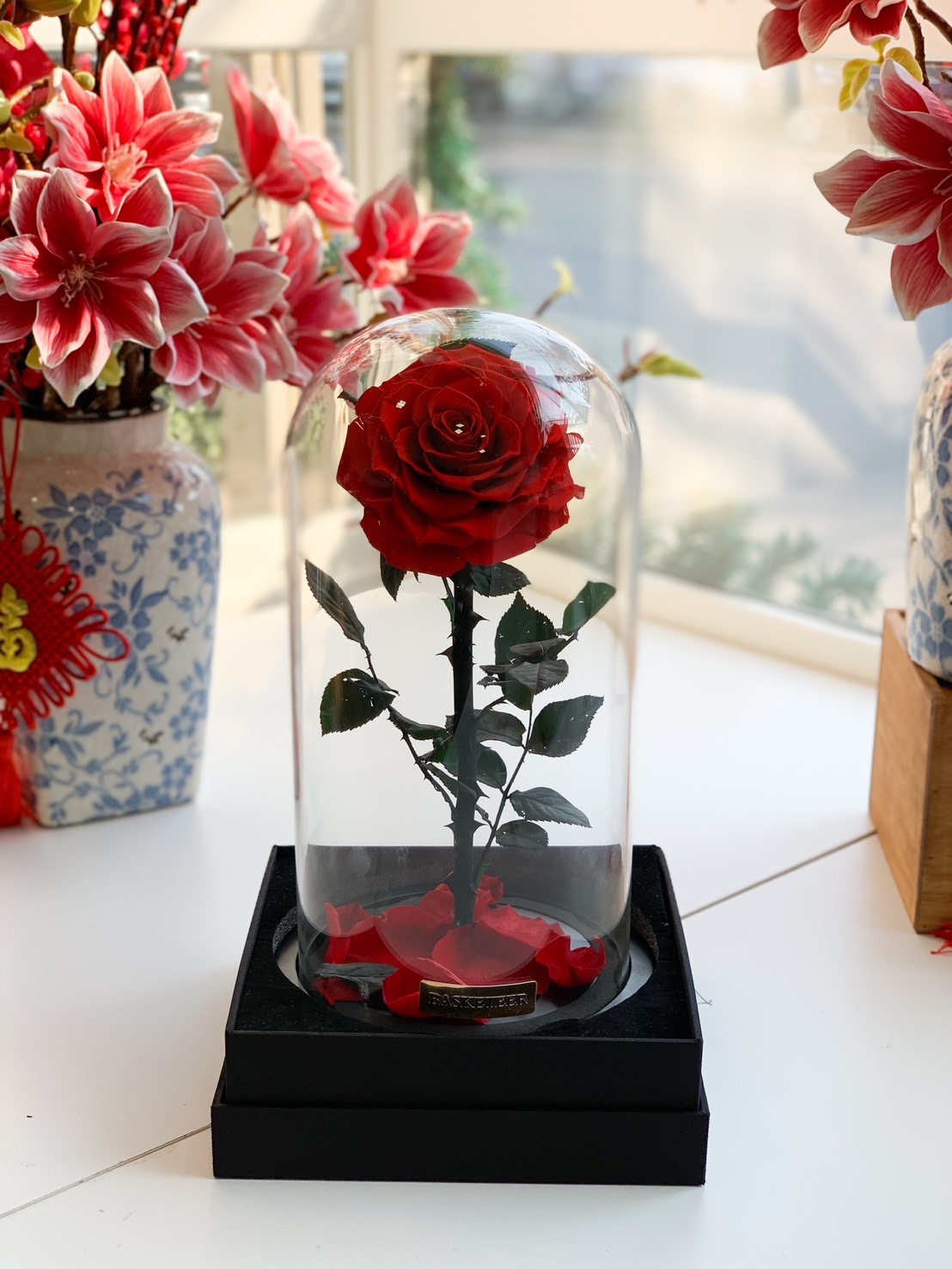 A luxurious preserved red rose in a glass dome, elegantly displayed on a black base with scattered rose petals – a timeless romantic gift.
