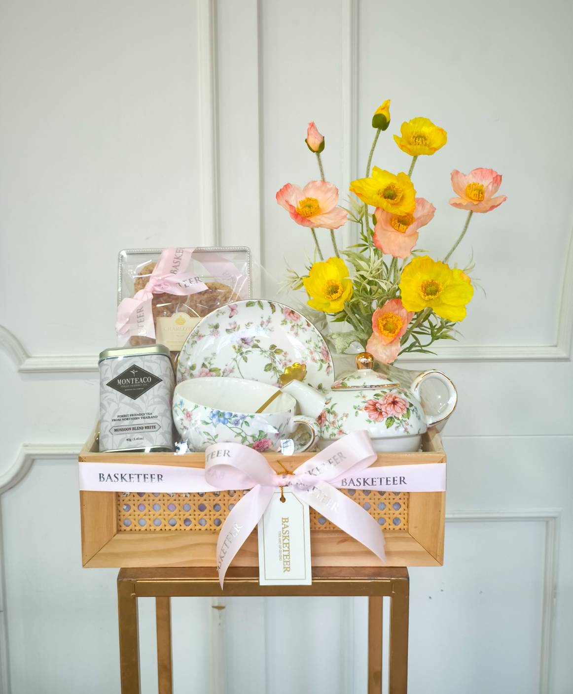 Elegant tea set gift box with floral porcelain teapot, cups, saucers, premium tea, biscuits, and fresh yellow-pink poppy flowers.