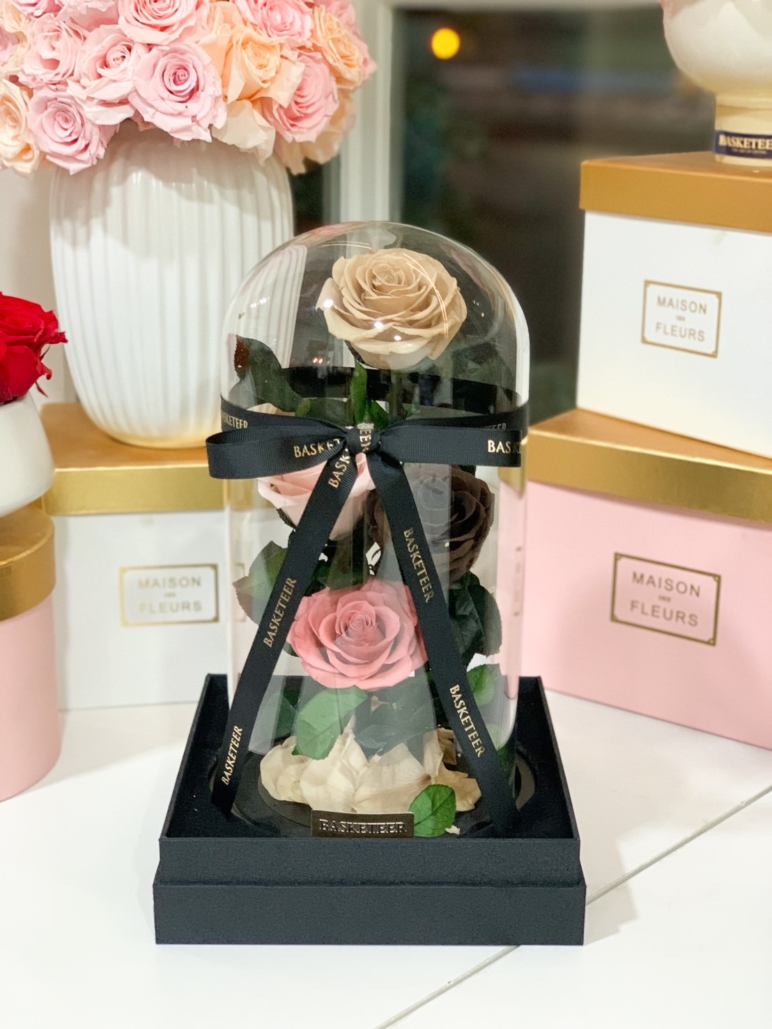 A luxury preserved rose arrangement with beige and pink roses in a glass dome, finished with a Basketeer ribbon and black gift base.