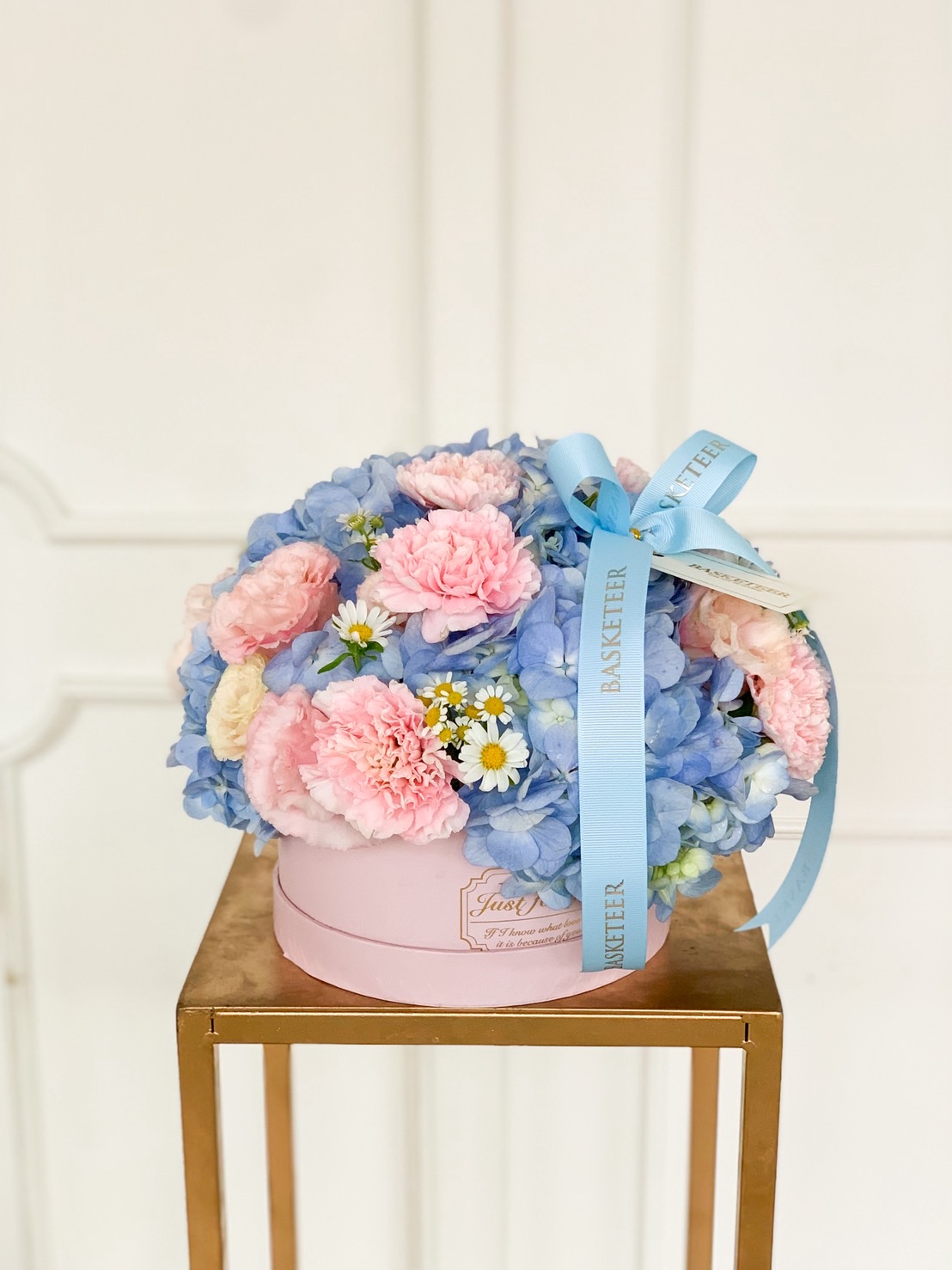 Pastel floral hatbox with blue hydrangeas, pink carnations, and daisy accents tied with a blue ribbon