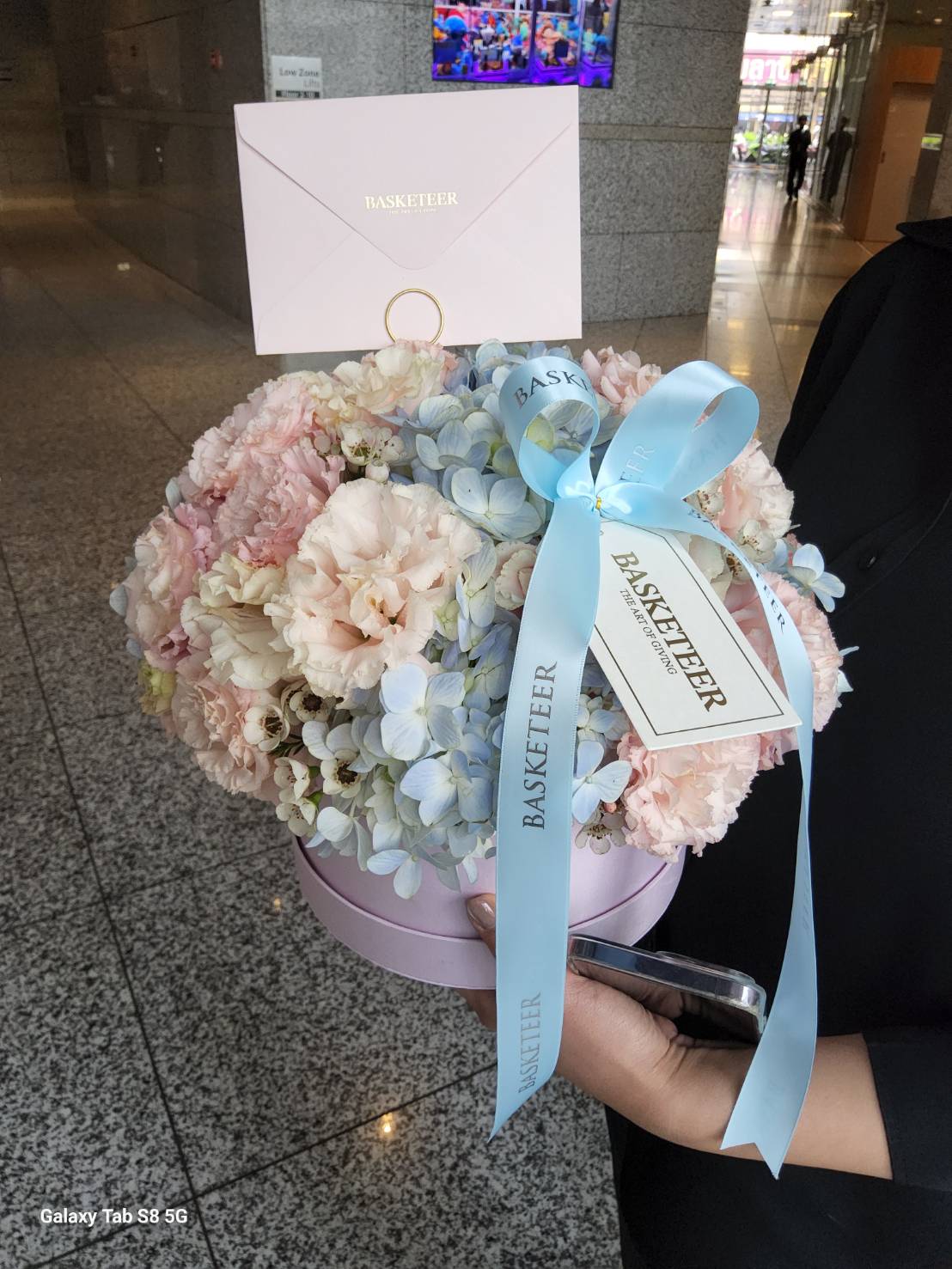 A luxurious pastel bloom box with hydrangeas, pink carnations, and delicate florals, adorned with a blue satin ribbon and a personalized greeting card, elegantly presented in a soft pink round box.