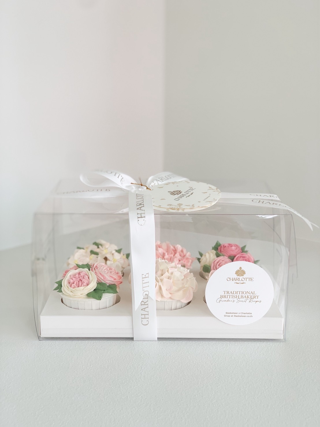 Elegant boxed set of floral buttercream cupcakes in pink and ivory, tied with a Charlotte ribbon for gifting.