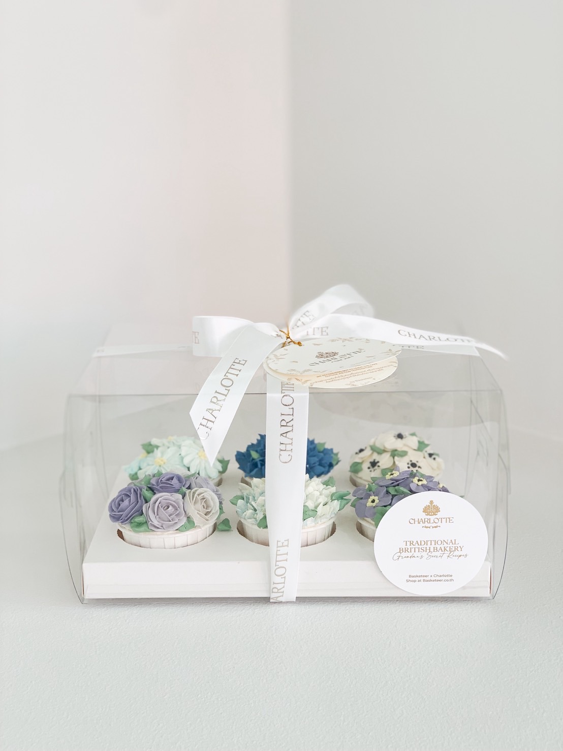 Boxed set of cupcakes decorated with blue and white buttercream flowers, tied with Charlotte ribbon.