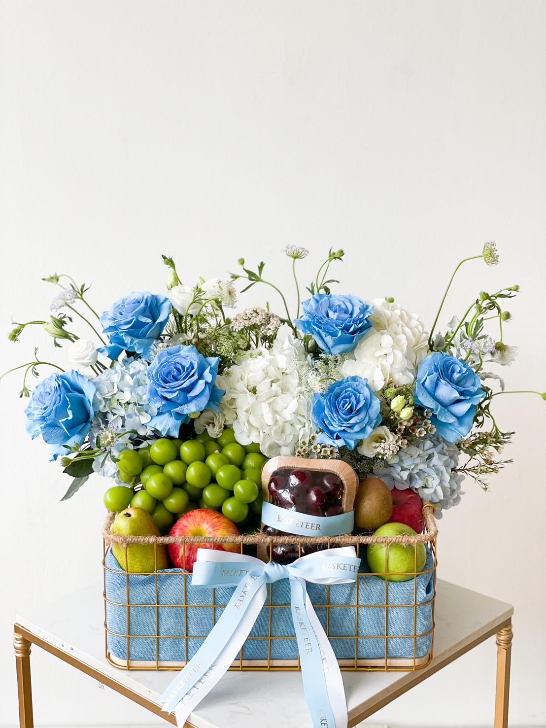 The Rose & Orchard Elegance Basket, filled with assorted fruits including green grapes, apples, and kiwi, is adorned with blue roses, hydrangeas, and other white flowers. Light blue ribbons are tied around the basket, which sits on a small white table against a plain background.