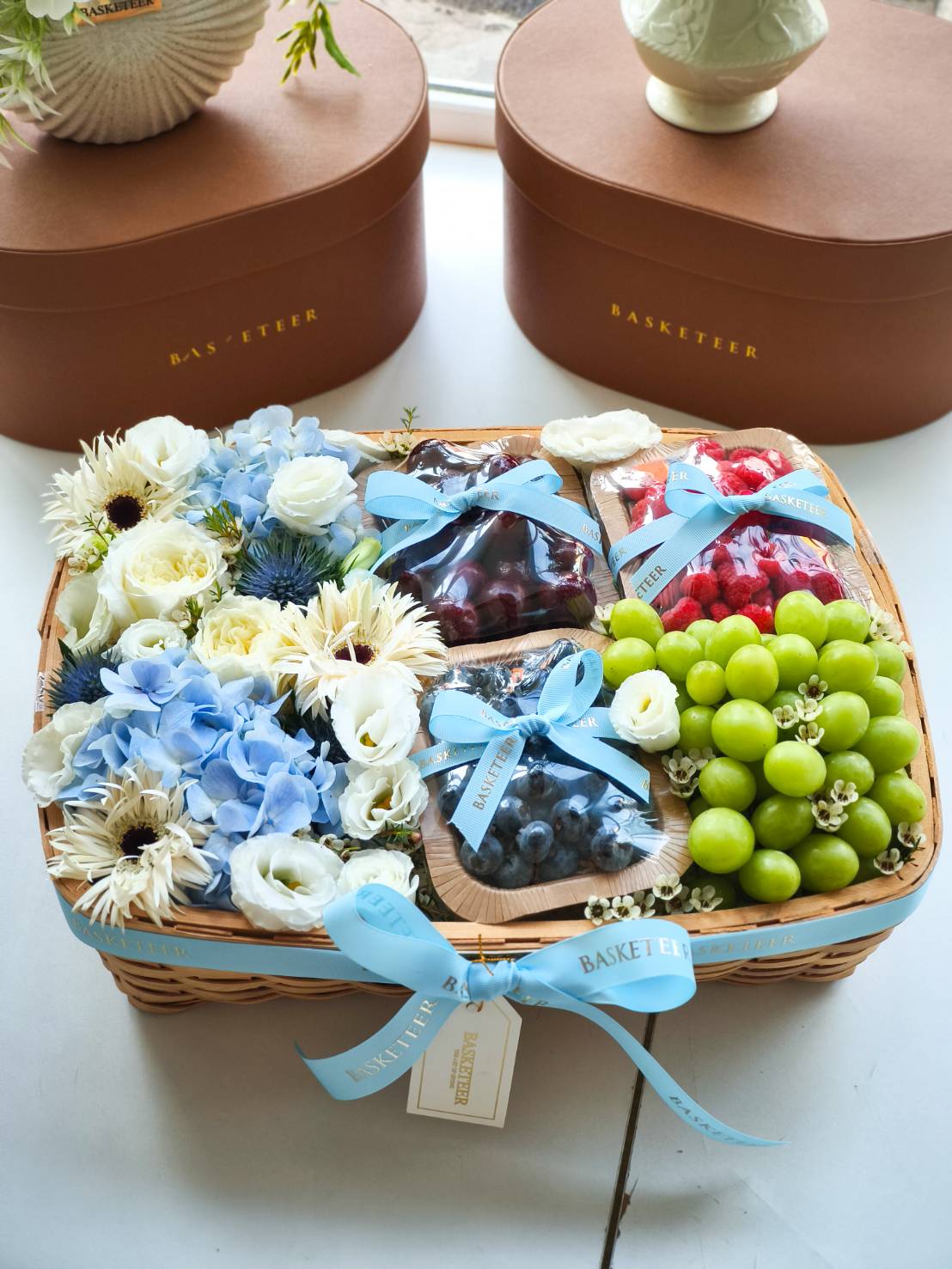 Luxury fruit basket with green grapes, blueberries, cherries, raspberries, decorated with white and blue flowers and a blue satin ribbon.