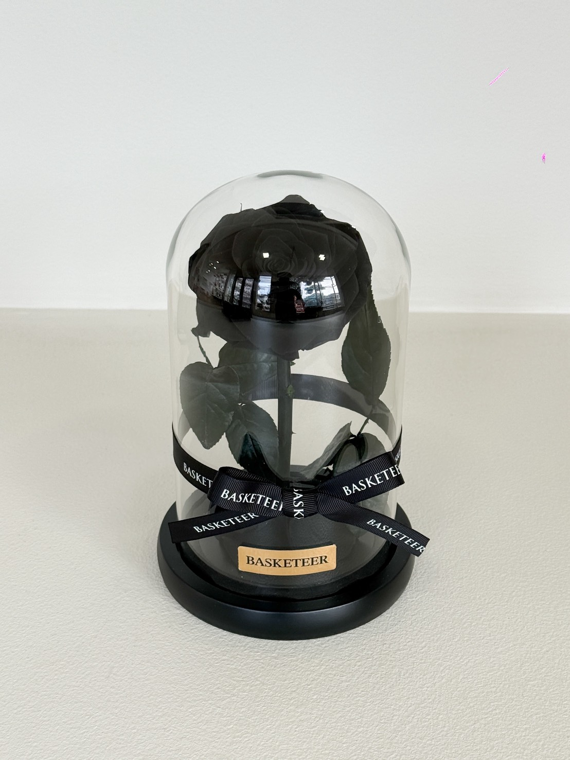 The Valentine’s Eternal Black Rose Dome features a single preserved black rose with green leaves under a glass dome on a black base, adorned with black BASKETEER ribbons and a matching gold plaque.