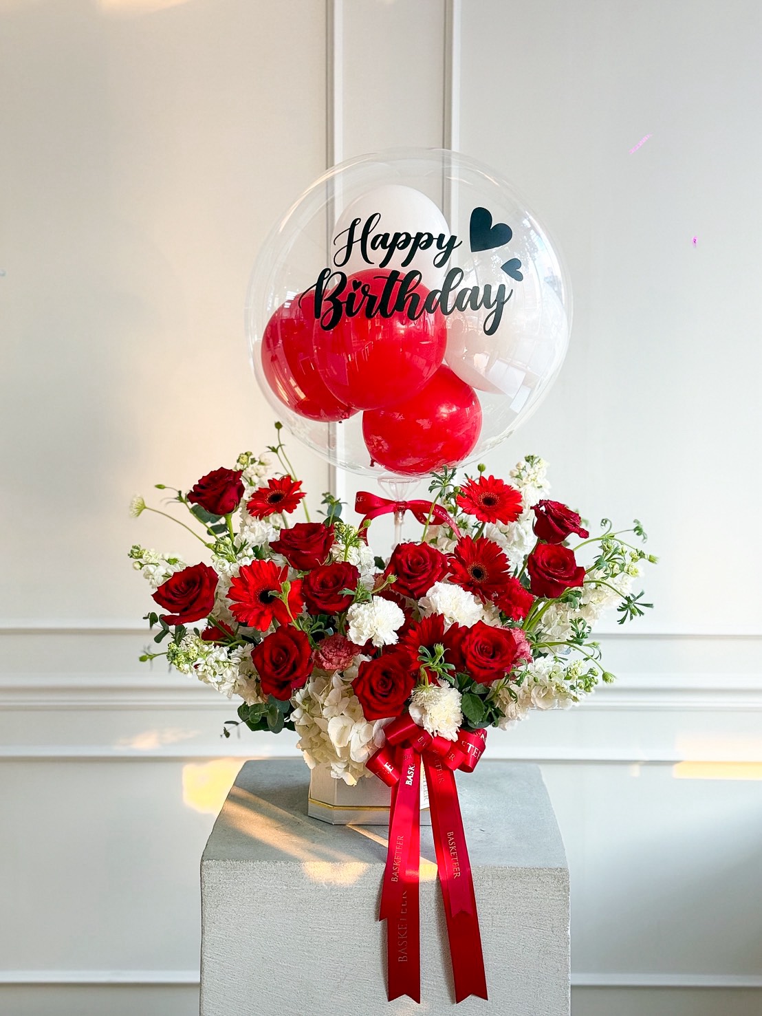 A Red Radiance Flowers with Balloon Gift features red and white blooms with a red ribbon on a pedestal. Above, a clear balloon filled with red balloons reads 